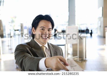 Businessman checking in at airport, receiving boarding pass from check-in attendant, smiling, portrait, view from behind check-in desk (differential focus)