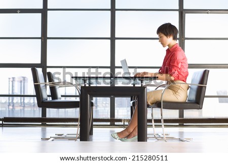 Young woman sitting at table by window, using laptop, side view