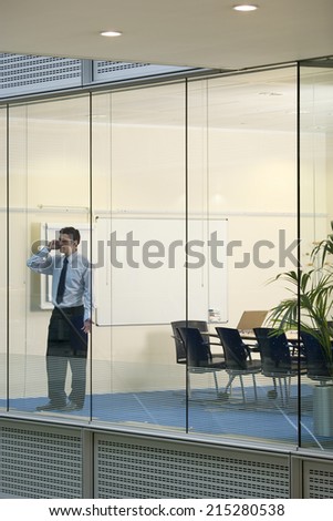 Solitary businessman standing in boardroom, using mobile phone, view through large office window