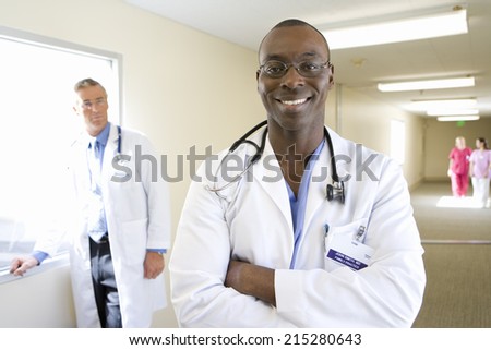 Young male doctor standing in hospital corridor, smiling, mature male doctor in background, portrait