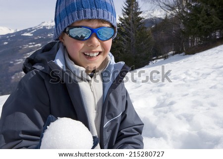 Boy wearing woolen hat and sunglasses in snow field, holding snow ball, smiling, portrait