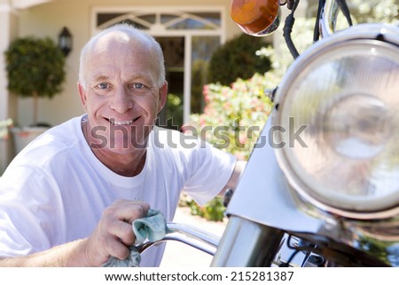 Senior man polishing motorbike on driveway, crouching down, smiling, close-up, portrait