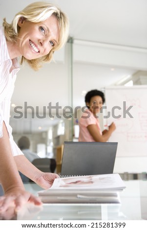 Two businesswomen in meeting room, one looking at book on table, one using flipchart, smiling, portrait (differential focus)