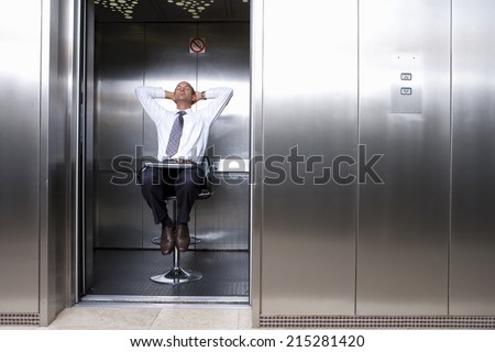 Mature businessman on stool in lift, laptop on lap, leaning back with head in hands