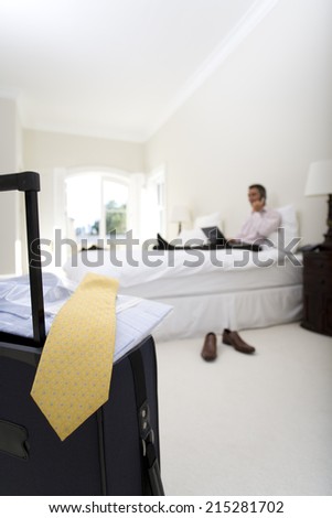 Mature businessman using laptop and mobile phone on bed, suitcase in foreground