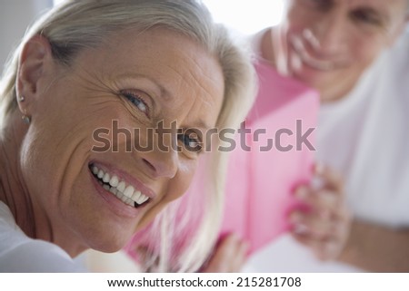 Mature man giving wife pink birthday gift, focus on woman looking over shoulder in foreground, smiling, close-up, portrait