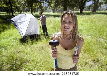 Young couple assembling dome tent on camping trip in woodland clearing, focus on woman holding tent peg and mallet in foreground, smiling, portrait