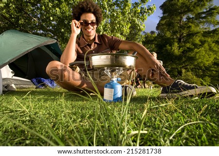 Young man, in sunglasses, cooking food on gas camping stove, smiling, front view, portrait (surface level)
