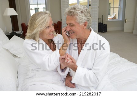 Mature couple sitting on bed, woman holding mobile phone by man\'s ear, smiling