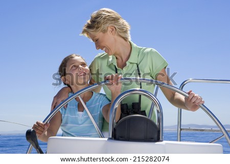 Mother and daughter (8-10) standing at helm of sailing boat out at sea, steering, smiling at one another, front view