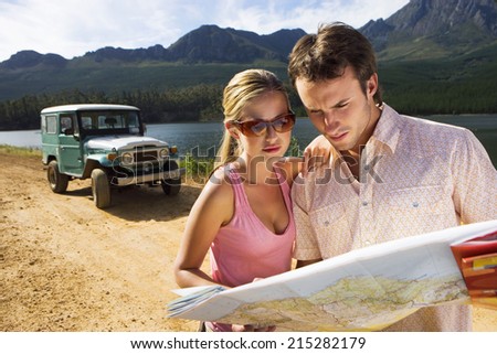 Young couple standing near parked jeep on dirt track beside lake, consulting map