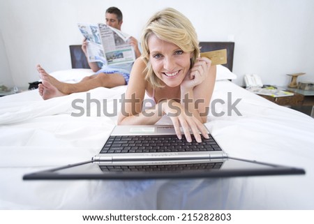 Woman using laptop on bed, smiling, portrait, man reading newspaper in background