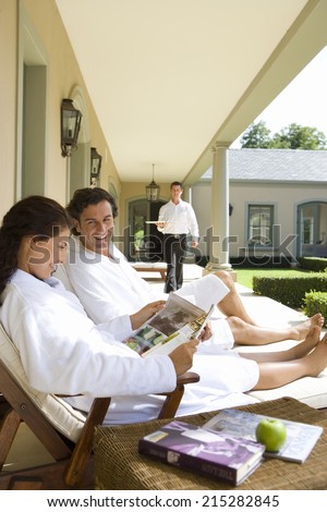 Couple wearing white bath robes on deck chairs, man smiling at woman holding magazine, waiter standing in background