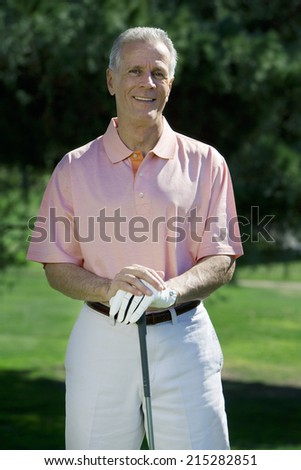 Mature man, in pink short-sleeved shirt and golf glove, standing on golf course, leaning on golf club, smiling, front view, portrait
