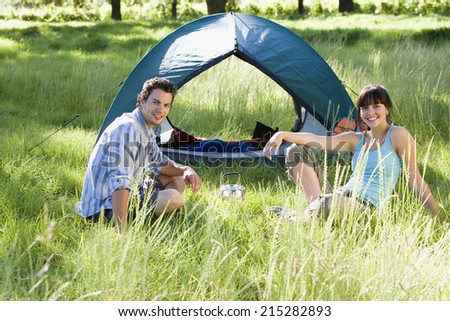 Young couple sitting near dome tent on camping trip in woodland clearing, smiling, side view, portrait