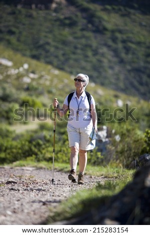 Mature woman hiking on mountain trail, carrying rucksack, using hiking pole, smiling, front view