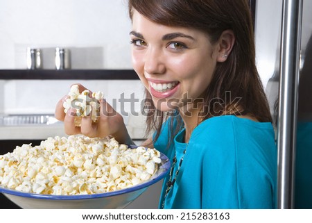 Young woman standing by fridge with bowl of popcorn, smiling, portrait, close-up