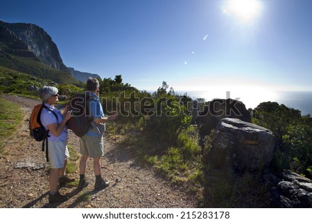 Mature couple hiking on mountain trail, looking at Atlantic Ocean horizon in bright sunlight, side view (lens flare)