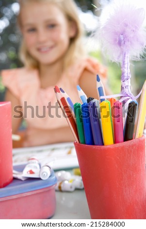 Girl (6-8) sitting at table with paints, smiling, portrait (focus on coloured markers in foreground)