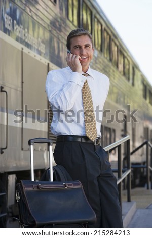 Businessman, with luggage, standing on railway platform beside stationary train, using mobile phone, portrait