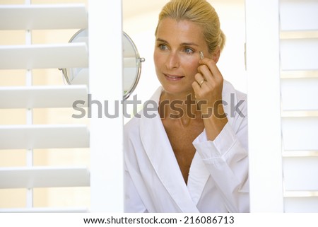 Woman plucking eyebrows by mirror, smiling, view through shutters