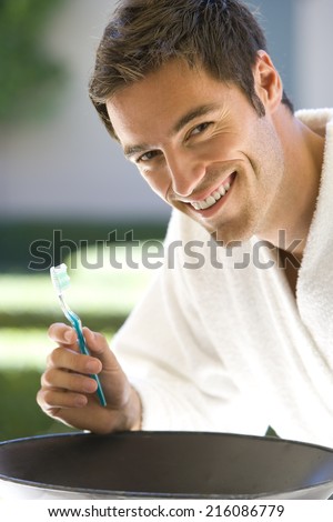 Young man in bathrobe brushing teeth, smiling, portrait