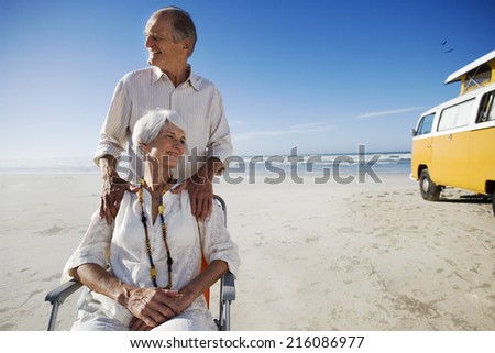 Senior couple on beach by camper van, man behind woman in chair, smiling