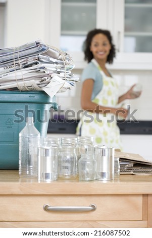 Glass jars and cans by recycling bin with newspapers in kitchen, woman in background