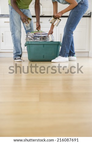 Couple, heads cropped, indoors, placing tins and jar into green recycling box with newspapers and magazines,