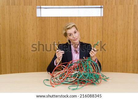 Businesswoman at table with bunch of tangled cable leads, holding up two ends, portrait