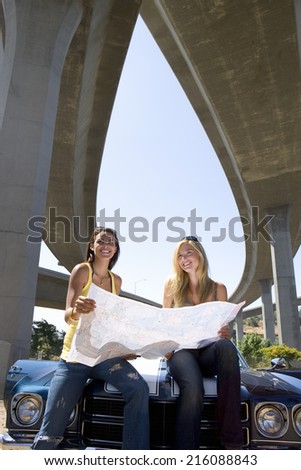 Two young women on bonnet of car looking at road map beneath overpass, smiling, portrait, low angle view