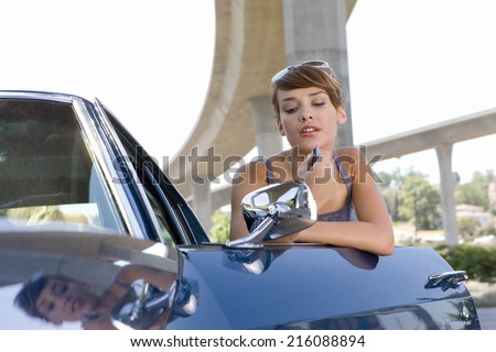 Young woman leaning on open door of car beneath overpass, looking in rear view mirror, low angle view