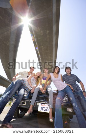 Medium group of friends on bonnet of car beneath overpass, smiling, low angle view (sun flare)