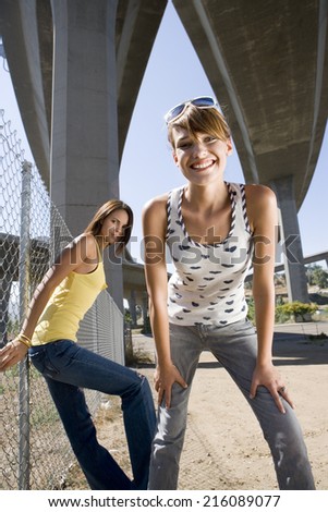 Young woman beneath overpasses, friend by fence, smiling, portrait, low angle view