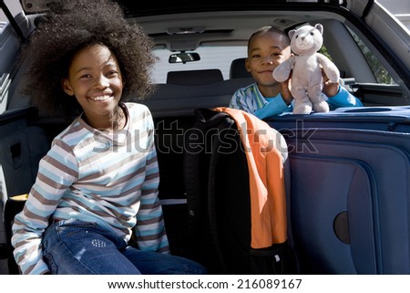 Brother and sister (6-10) in back of car with luggage, boy holding toy, smiling, portrait