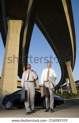 Two businessmen by car with briefcases beneath overpasses, low angle view