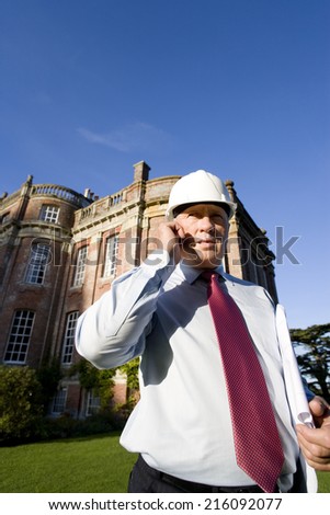 Businessman in hardhat using mobile phone by manor house, low angle view