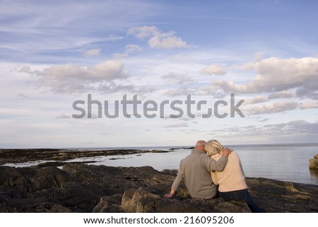 Senior couple on rocks looking out to sea, man with arm around woman, rear view