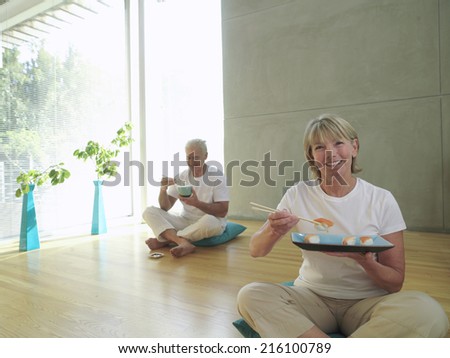 Mature couple on cushions, woman eating sushi with chopsticks, smiling, portrait