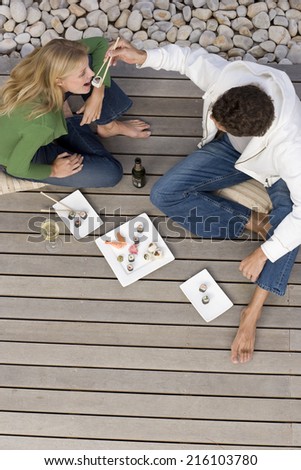 Young couple on decking, man feeding woman sushi with chopsticks, elevated view