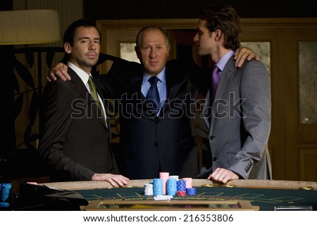 Mature man with arms around young men by poker table, portrait