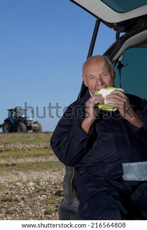 Farmer eating sandwich in back of car with tractor in background