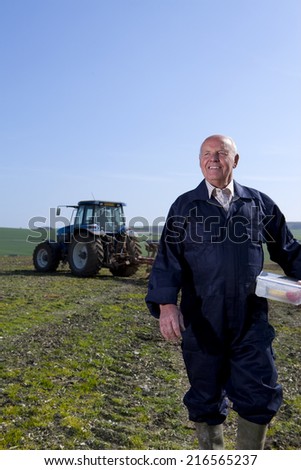 Smiling farmer holding lunchbox in field with tractor and plough in background