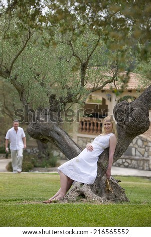 Smiling woman leaning on olive tree trunk with man in background