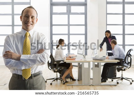 Smiling businessman standing with arms crossed and co-workers working in background