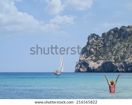 Woman wading in ocean with arms raised and sailboat in distance