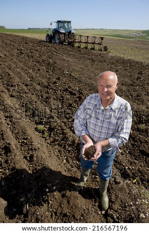 Farmer cupping soil in ploughed field with tractor and plough in background