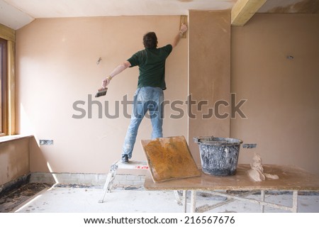 Man on step stool plastering wall in house under construction