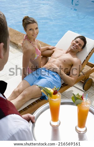 Waiter serving tropical drinks to couple on lounge chair at poolside