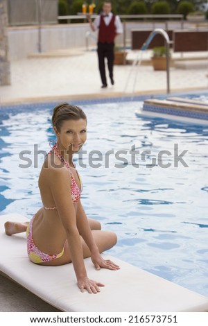 Smiling woman in bikini sitting at poolside with waiter in background